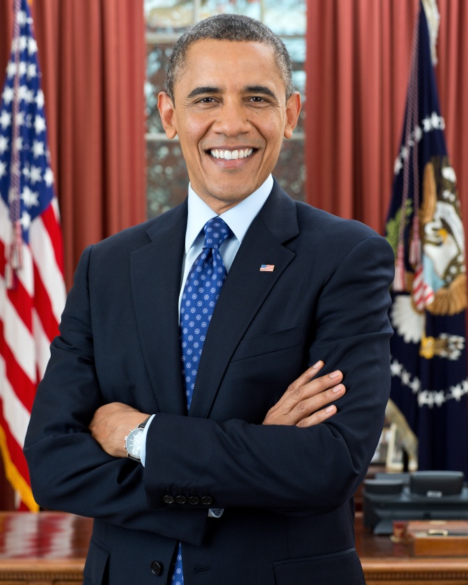 Barack Obama in einem blauen Anzug mit Krawatte. Er steht lächelnd mit verschränkten Armen vor der amerikanischen Flagge.