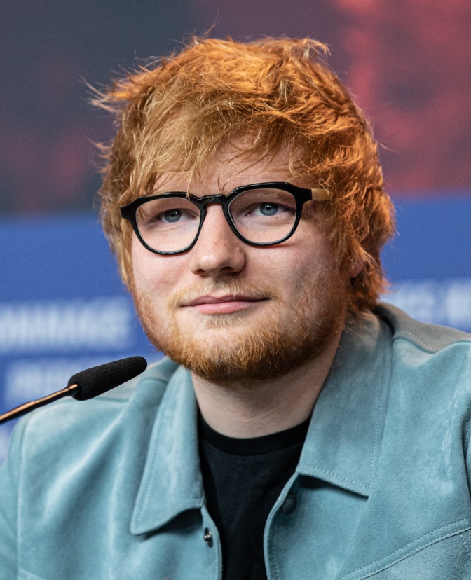 Ed Sheeran mit roten Haaren und rotem Bart. Er trägt eine massive, schwarze Brille und ein hellblaues Hemd über einem T-Shirt.