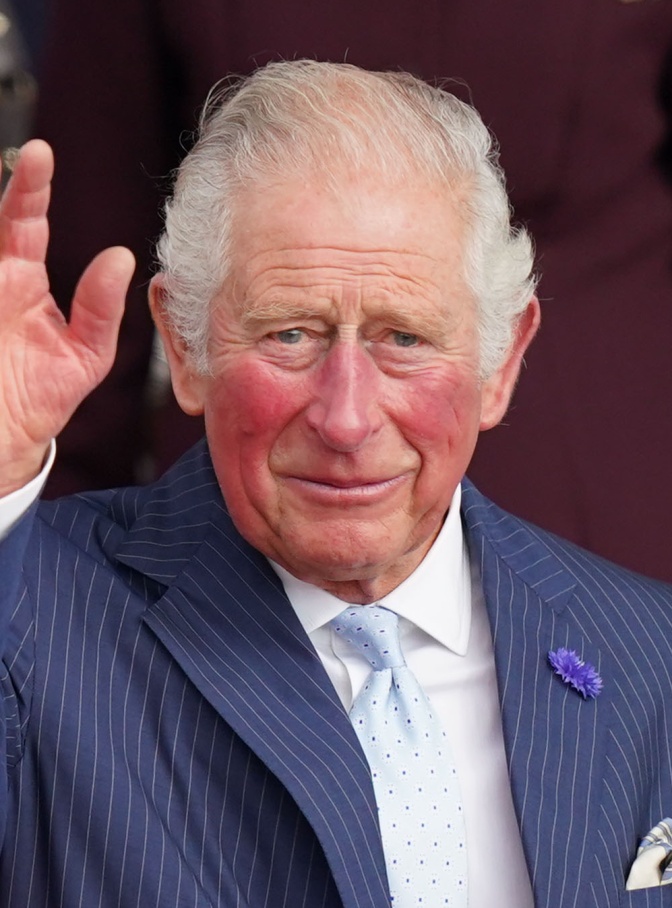 König Charles in einem blauen Anzug und hellblauer Krawatte. Er winkt mit erhobener Hand. Er hat weiße, über die Halbglatze gekämmte Haare und sehr rote Wangen.