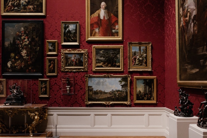 Mehrere Gemälde in goldenen Rahmen hängen über- und nebeneinander an einer Wand im Museum.