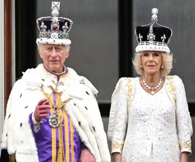 Charles und Camilla tragen ihre Kronen und stehen nebeneinander. Charles trägt einen Hermelinmantel, Camilla ein weißes, besticktes Kleid.