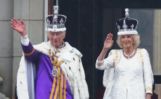 Charles und Camilla mit Kronen auf dem Kopf und in festlicher Kleidung. Sie winken vom Balkon aus.
