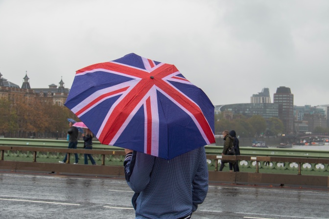 Ein Mann, von hinten fotografiert, im Regen, unter einem Regenschirm mit dem Motiv der britischen Flagge.