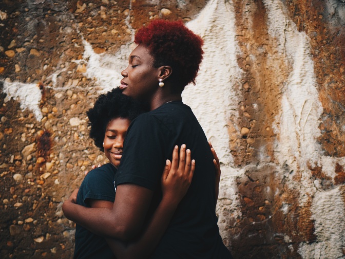 Eine schwarze Frau umarmt ihre Tochter. Beide haben krauses Haar und tragen schwarze T-Shirts. Sie haben die Augen geschlossen.