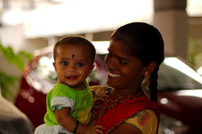 Eine indische Mutter mit dunkler Haut und geflochtetenen schwarzen Haaren. Sie trägt einen Sari und hat ein Bindi auf der Stirn. Sie hält ein Kleinkind im Arm, beide lächeln.