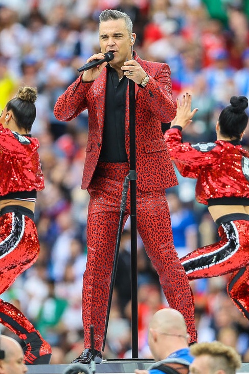 Robbie Williams in einem roten Anzug auf der Bühne. Er singt und hält sich am Mikrofonständer fest. Im Hintergrund sieht man eine große Menschenmenge.