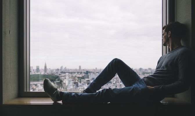 Ein Mann sitzt innen im Raum auf einer Fensterbank. Vor dem Fenster grauer Himmel und eine Großstadt.