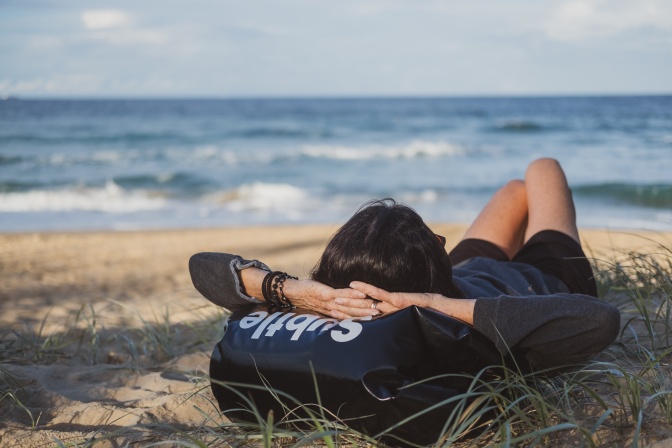 Eine Frau liegt am Strand im Sand auf dem Rücken. Unter dem Kopf liegt ein Beutel, die Arme sind hinter dem Kopf verschränkt.