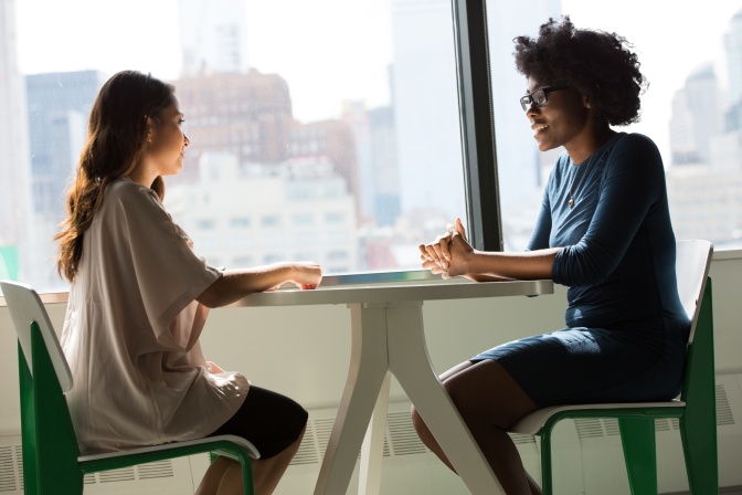2 Frauen sitzen sich gegenüber an einem runden Tisch und unterhalten sich. Eine der beiden ist schwarz und hat einen Afro. Sie trägt Brille. Die andere der beiden ist BiPOC und hat gewellte, lange Haare.