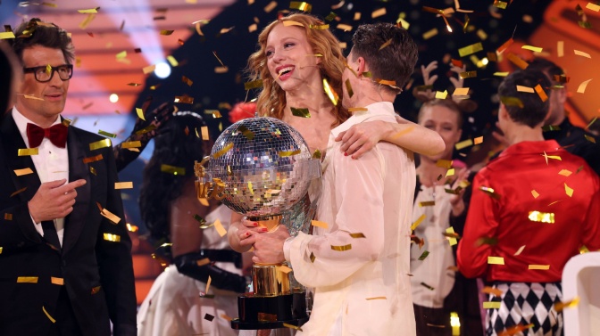 Anna Ermakova und ihr Tanzpartner Valentin Lusin stehen Arm in Arm unter einem Regen aus goldenem Konfetti. Sie hält den Siegerpokal in der Hand. Hinter den beiden stehen Menschen und klatschen.