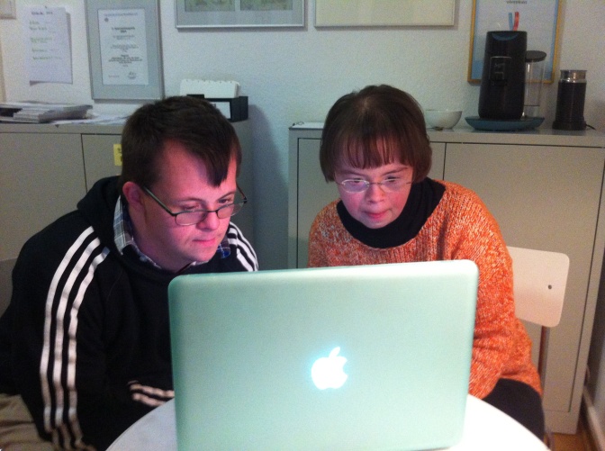 Ein Mann und eine Frau mit Down-Syndrom sitzen vor einem Laptop und lesen gemeinsam einen Text.
