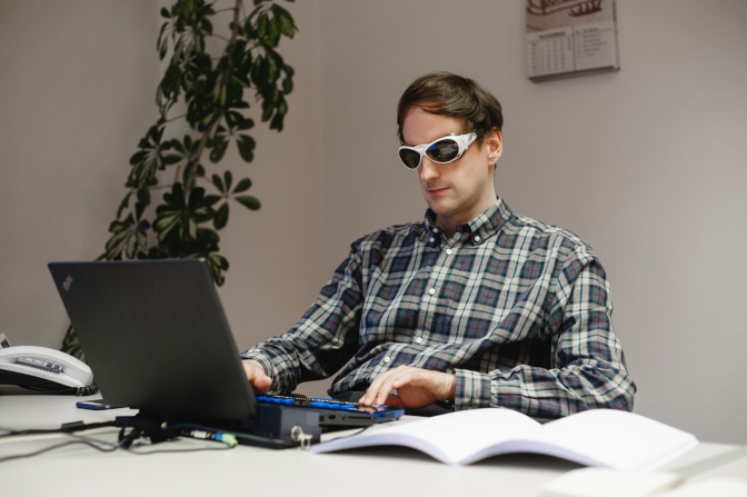 Ein blinder Mann mit dunkler Brille sitzt an einem Laptop. Neben ihm liegt ein aufgeschlagenes Buch.