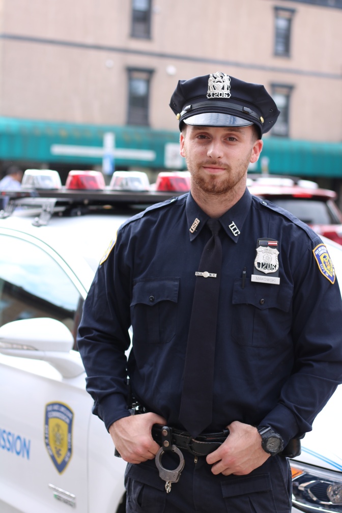 Ein Mann steht in Uniform der amerikanischen Polizei an ein Auto gelehnt. Die Uniform ist dunkelblau, an der Brust hat er seine Dienstnummer angesteckt.