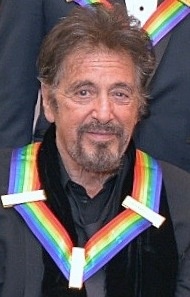 Al Pacino mit einer Kette aus regenbogenfarbenen Bändern um den Hals. Er hat graue Haare und einen rauen Vollbart und schaut in die Kamera.