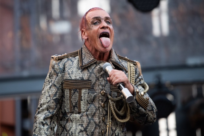 Till Lindemann steht mit herausgestreckter Zunge auf der Bühne. Er hält ein Mikro in der Hand und trägt eine Jacke mit Schlangenmuster.