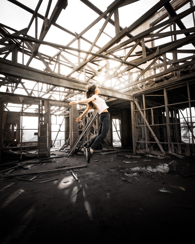 Eine Frau in einer Industriehalle mit Metallstreben und einer offenen Decke. Sie steht mitten im Raum und springt in die Luft.