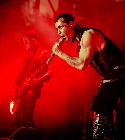 Till Lindemann auf der Bühne. Er trägt einen roten Kragen aus Federn und singt ein ein Mikro. Die Bühne ist rot beleuchtet.