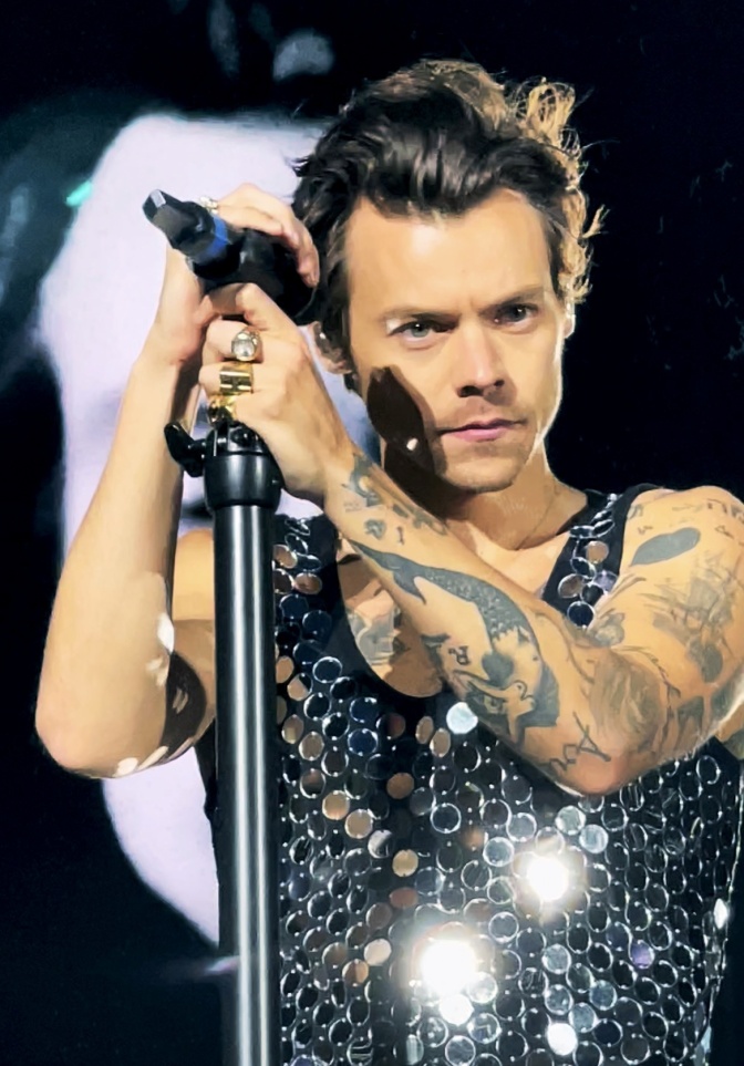 Harry Styles in einem Glitzershirt am Mikrofon. Er hat mehrere Tattoo und kurze, braune Haare.