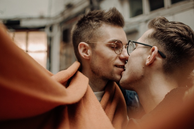 Zwei Männer küssen sich. Um sie herum weht ein orangefarbener Schal, im Vordergrund des Bildes ist es unscharf.