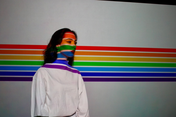 Eine Frau im weißen Sweatshirt steht vor einer Wand, an die ein Regenbogen projiziert wird. Die Projektion läuft auch über das Gesicht der Frau.