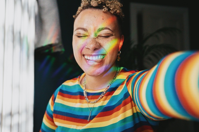 Eine Person of Color in einem Shirt in Regenbogenfarben. Sie hat die Augen geschlossen und auf ihrem Gesicht sieht man Licht, das sich in Regenbogenfarben bricht.