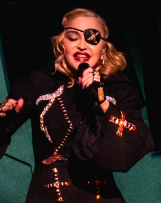 Madonna in einem schwarzen Bühnenoutfit mit Augenklappe und starkem Make up. Sie singt in ein Mikro.