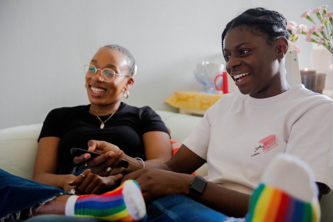 2 schwarze Personen sitzen lächelnd zusammen auf einer Couch. Beide tragen regenbogenfarbene Socken.