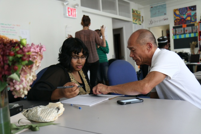 Ein älterer asiatischer Mann und eine junge, schwarze Frau sitzen zusammen an einem Tisch. Er beugt sich zu ihr herüber und zeigt auf ihre Notizen.