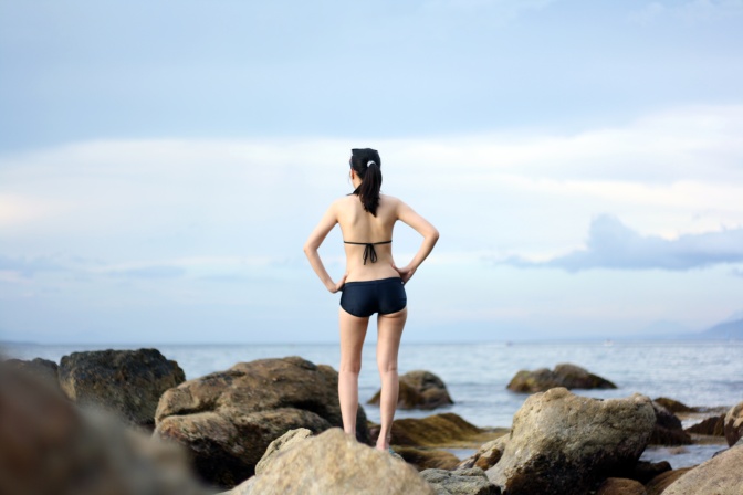 Eine Frau steht mit in die Hüfte gestemmten Armen im Bikini auf einem Felsen. Man sieht sie von hinten.