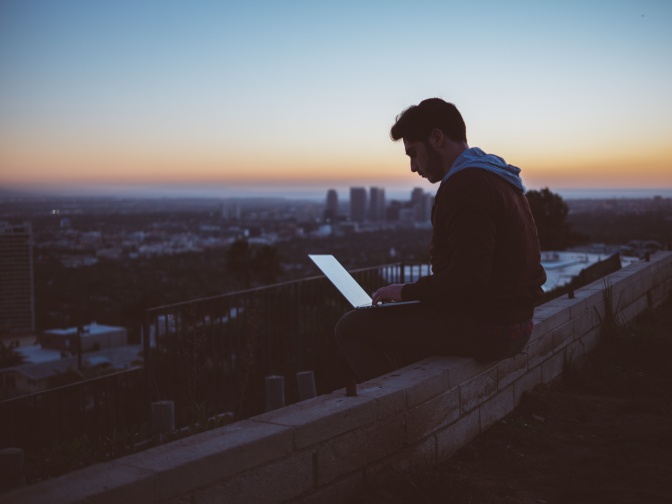 Ein Mann mit Schiebermütze sitzt in der Dämmerung im Freien auf einer Mauer und arbeitet am Laptop.