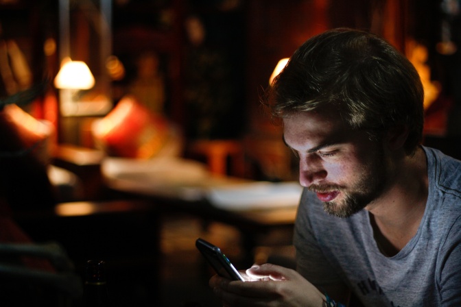 Ein Mann sitzt in einer Bar. Das Licht ist gedämpft, er scrollt am Handy und wird vom Display angestrahlt.