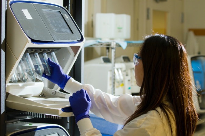 Eine Frau im weißen Kittel und mit blauen Plastikhandschuhen. Sie trägt eine Schutzbrille und arbeitet in einem Labor an einem technischen Gerät.