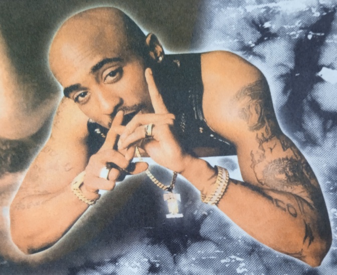 Tupac Shakur ist BiPoc, hat eine Glatze und viele Tattooss auf den Armen. Er hat den Kopf auf die Hände gestützt und schaut in die Kamera.