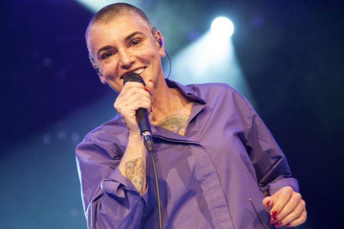 Sinéad O’Connor in einer lilanen Bluse mit sehr kurz rasierten Haaren. Sie steht auf der Bühne und hält lächelnd ein Mikro in der Hand.