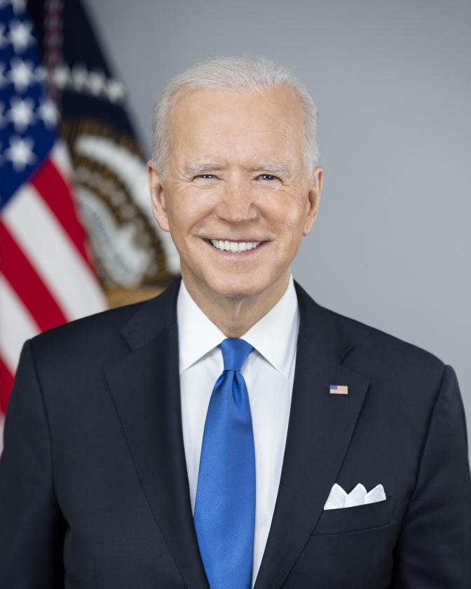 Joe Biden mit weißen Haaren in Anzug und Krawatte. Er steht lächelnd vor einer amerikanischen Flagge.