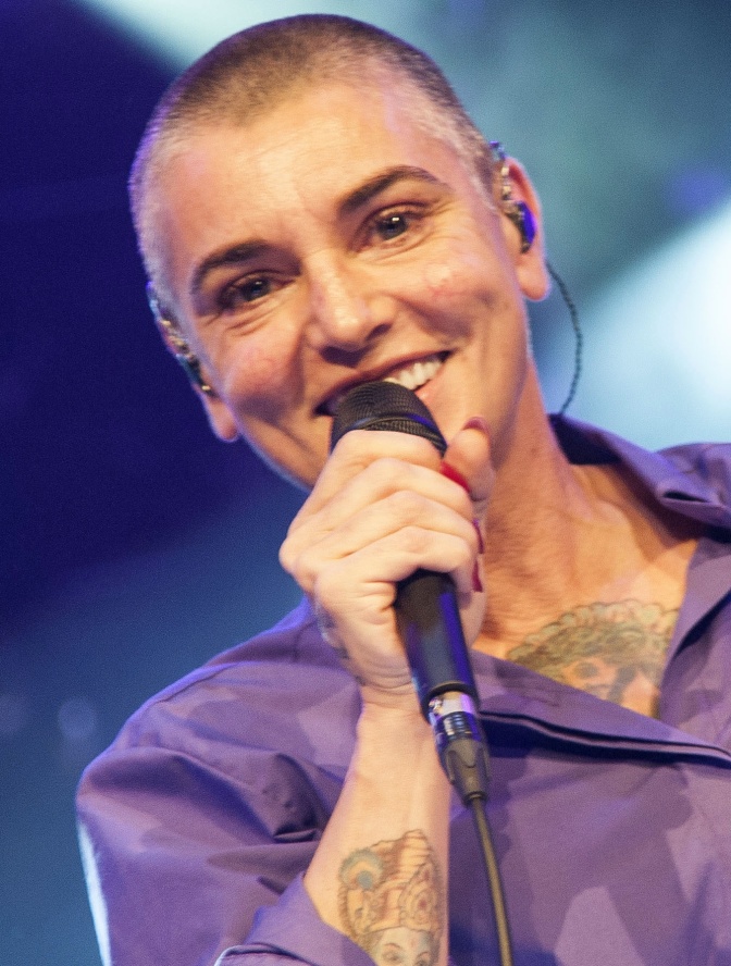 Sinéad O'Connor mit sehr kurz rasierten Haaren in einer Lianen Bluse. Sie singt lächelnd in ein Mikro.