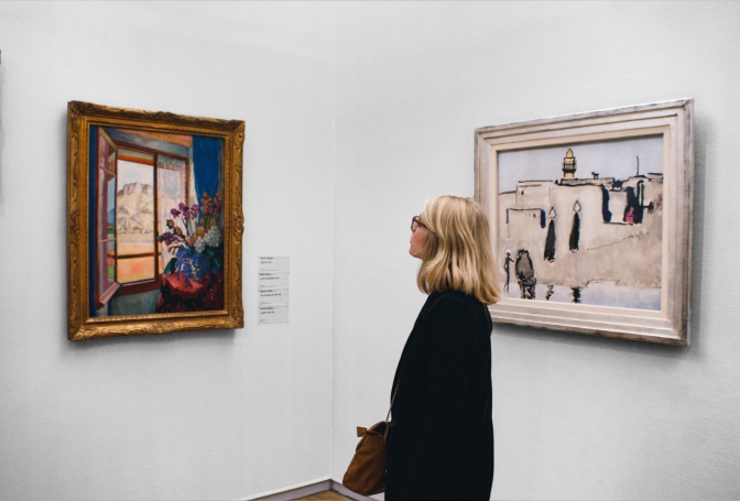 Eine schwarz gekleidete Frau steht im Museum vor einem Gemälde und betrachtet es. Hinter ihr hängt ein weiteres Gemälde an der Wand.