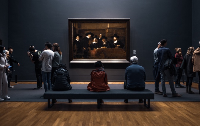 Menschen in einem Museum. 3 Menschen sitzen auf einer Bank mit dem Rücken zur Kamera und betrachten ein Gemälde. Andere stehen vor anderen Bildern oder machen Selfies mit sich und den Bildern.