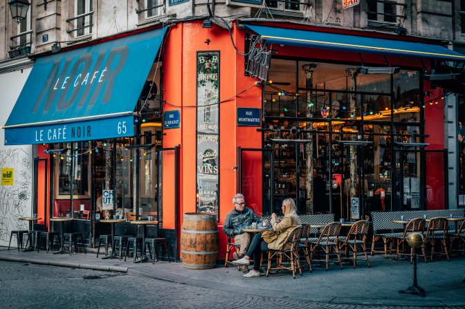 Ein Mann und eine Frau sitzen in Jacken im Freien an dem Tisch eines Straßencafés. Das Café ist in einem in kräftigem Orange gestrichenen Eckhaus.