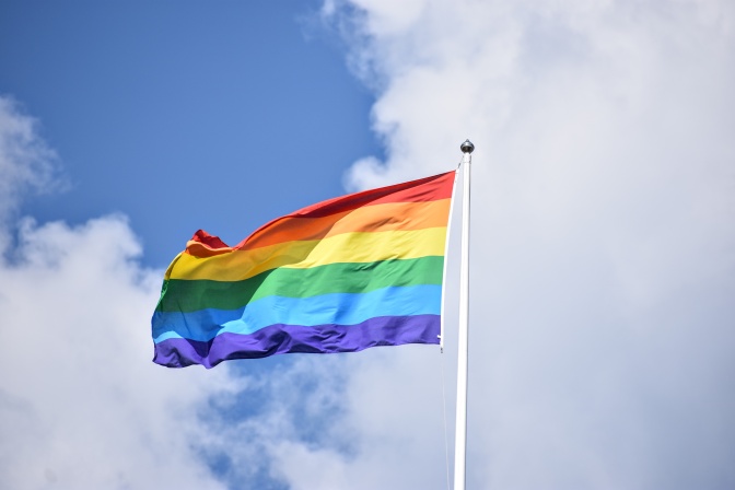 Eine Regenbogenflagge mit Streifen in rot, orange, gelb, grün blau und lila vor leicht bewölktem Himmel.
