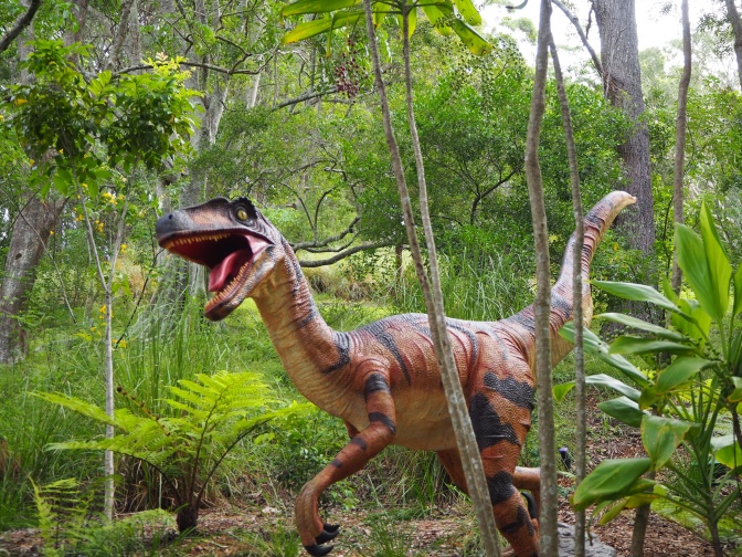 Ein fleischfressender Dinosaurier mit offenem Maul und kleinen Ärmchen rennt durch einen Wald.