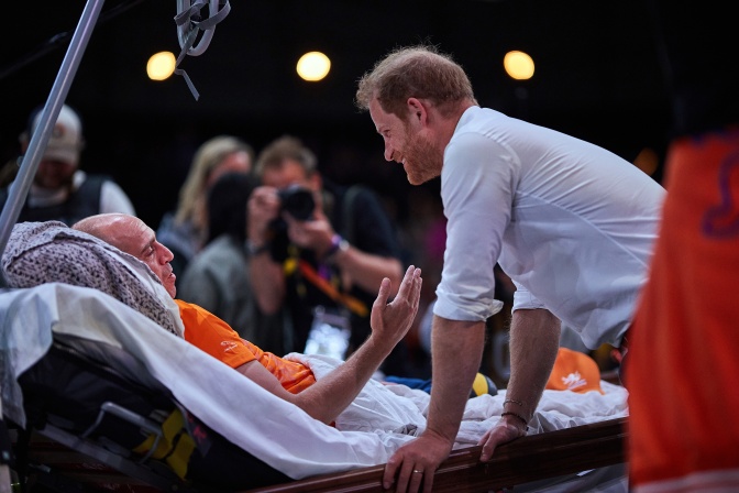 Prinz Harry spricht mit einem Mann, der auf einer Trage liegt. Der Mann gestikuliert beim Sprechen mit der Hand. Beide werden im Gespräch fotografiert und mit Strahlern angeleuchtet.
