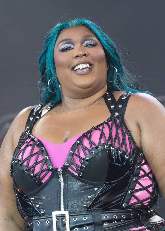 Lizzo ist eine schwarze Frau mit grünen Haaren. Sie trägt ein Bühnenkostüm in schwarz und pink.