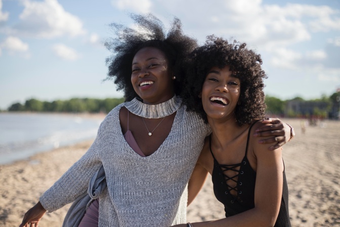 2 schwarze Frauen laufen lachend und Arm in Arm einen Strand entlang.