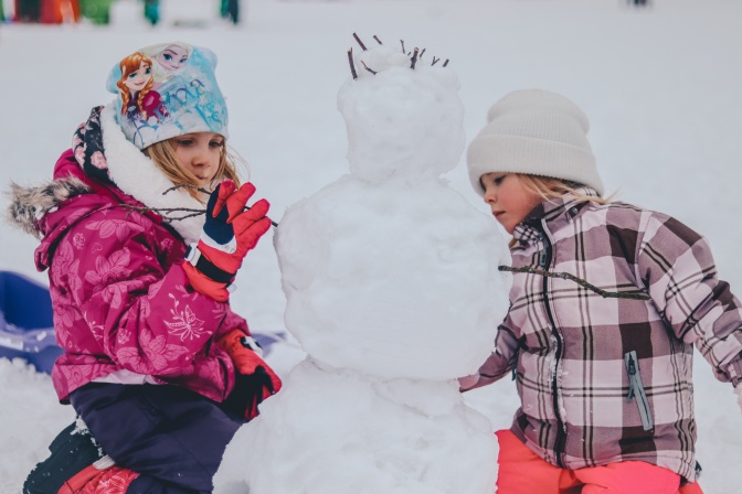 Zwei kleine Mädchen in Winterleidung bauen zusammen einen Schneemann.