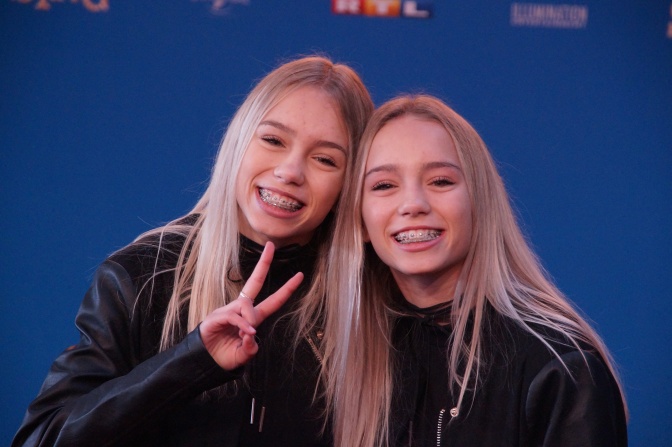 2 junge Frauen mit langen, glatten blonden Haaren und Zahnspangen. Sie tragen dunkle Basics und haben die Köpfe lächelnd aneinander gelehnt.