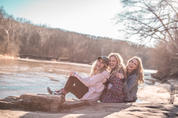Eine Gruppe von 3 jungen blonden, langhaarigen Frauen. Sie sitzen aneinander gelehnt auf dem Boden und lachen.