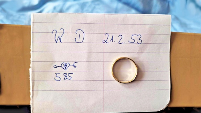 Ein schlichter, goldener Ehering liegt auf einem Zettel, auf dem in Handschrift die Gravur des Rings notiert ist: WD 21.3.1953