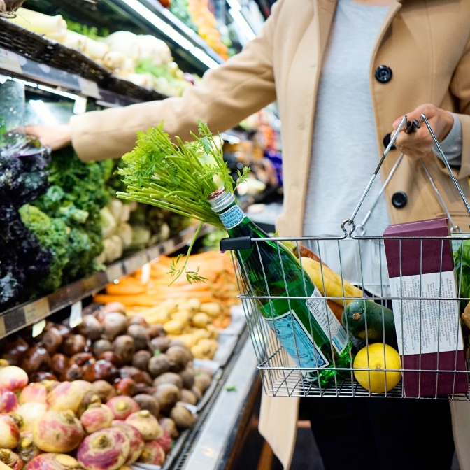 Eine Frau trägt einen Einkaufskorb am Arm und wählt Obst und Gemüse aus. Ihr Gesicht ist auf dem Foto nicht zu sehen.
