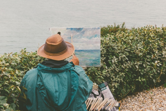 Ein Mann in Regenjacke und mit Hut sitzt am Wasser und malt auf einer Staffelei. Vor ihm eine blühende Hecke.